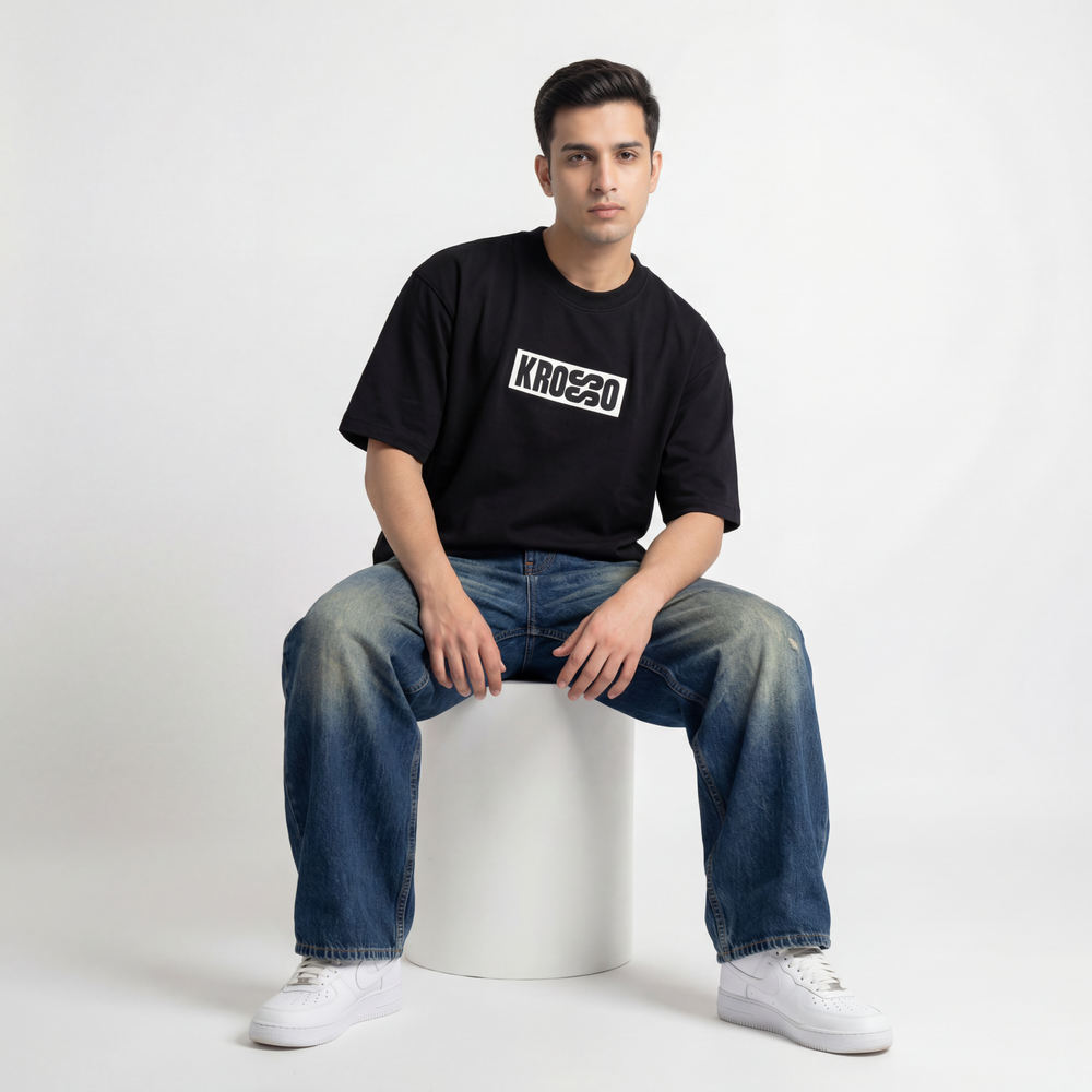 Front view of model sitting and wearing Krosso oversized black t-shirt with minimal logo print and oversized streetwear fit.